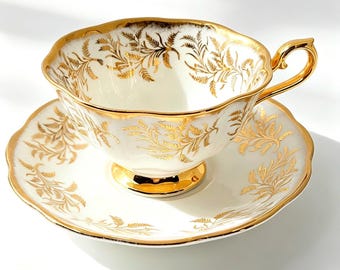 May include: A white porcelain teacup and saucer set featuring a scalloped edge and a delicate gold fern leaf pattern. The teacup has a gold-trimmed handle and a pedestal base, while the saucer displays a matching gold botanical design.