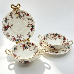 May include: A set of two cream-coloured porcelain bouillon cups and two matching saucers, featuring a delicate floral pattern in red and blue with brown vines. Each cup has two handles and gold trim. One saucer is displayed upright on a gold bow stand.