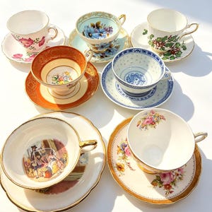 May include: A collection of seven ceramic teacups and saucers featuring diverse designs, including floral patterns, holly leaves, a blue and white dragon motif, and a historical scene with figures, all accented with gold trim and scalloped edges.