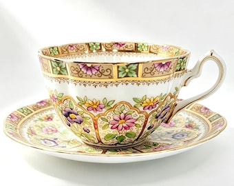 May include: A white porcelain teacup and saucer set featuring a decorative border of pink, yellow, and purple flowers with green leaves. The rim and handle are accented with gold trim, and the design includes intricate scrollwork patterns.