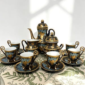 May include: A black and gold tea set with floral patterns. The set includes a teapot, creamer, sugar bowl, and six teacups with saucers. The teacups have ornate handles and sit on small feet. The set is displayed on a patterned tablecloth.