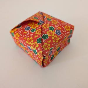 May include: A small, square origami box made from red paper with a floral pattern in yellow, green, and blue. The box is folded with precise edges and corners, showcasing the art of paper folding.