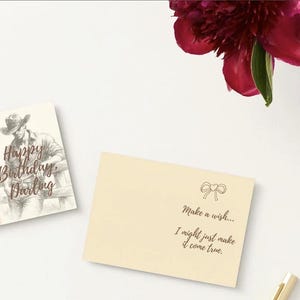 Könnte beinhalten: Zwei cremefarbene Grußkarten. Eine zeigt eine Cowboy-Illustration und den Text "Happy Birthday, Darling". Die andere Karte hat eine Schleifenillustration und den Text "Make a wish... I might just make it come true." Eine dunkelrote Blume ist sichtbar.