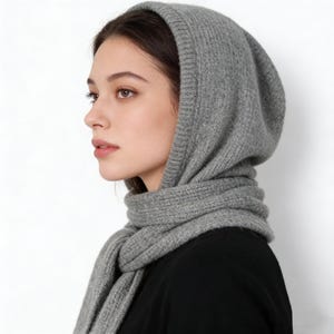 May include: A gray knit hood and scarf set. The hood is pulled up, framing the face, and the scarf is draped around the neck. The set is made of a soft, textured material, perfect for cold weather. The neutral color makes it versatile.