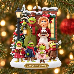 May include: A Muppets-themed Christmas ornament featuring Kermit the Frog, Miss Piggy, and four smaller frog characters in winter attire. The scene includes a brick wall, a street lamp, and a snow-covered base with the text The Garner Family.