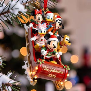 Puede incluir: Un adorno navideño que muestra a Mickey Mouse, Minnie Mouse, el Pato Donald, Daisy, Goofy y Pluto en un trineo rojo. El trineo está decorado con acebo y presenta el texto The Johnsons y el año 2026 en letras doradas.