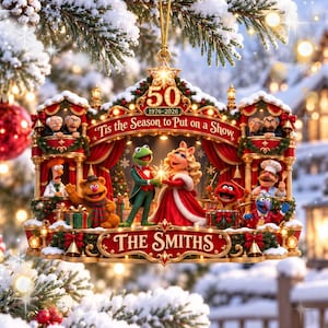 Könnte beinhalten: Ein Muppets-Weihnachtsanhänger mit Kermit der Frosch und Miss Piggy auf einer Bühne mit weiteren Figuren. Der Text lautet 50 1976-2026, Tis the Season to Put on a Show und THE SMITHS. Es ist ein mehrfarbiger Weihnachtsschmuck mit Goldrand.
