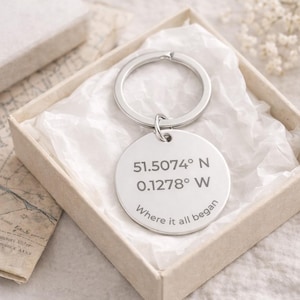 May include: A silver-coloured round keyring with a ring, presented in a beige box with white tissue paper. The keyring has engraved coordinates and the phrase "Where it all began."