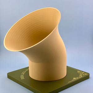 Op de afbeelding: Een beige, gebogen, trechtervormig object gemonteerd op een vierkante, olijfgroene basis. De basis is voorzien van goudkleurige decoratieve lijntekeningen van een kattengezicht en pootafdrukken, met de tekst "YOUR TEXT HERE" aan de zijkant.