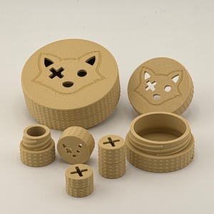 Puede incluir: Un conjunto de contenedores y componentes de color beige, impresos en 3D. El conjunto incluye cajas redondas con diseños de cara de zorro, piezas con tapa de rosca y piezas cilíndricas más pequeñas con detalles en forma de cruz. Las piezas tienen una superficie texturizada.