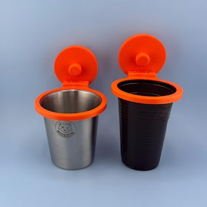 Może przedstawiać: Dwa uchwyty na kubki z jaskrawopomarańczowymi plastikowymi obręczami i przymocowanymi pokrywkami. Lewy uchwyt wykonany jest ze szczotkowanej stali nierdzewnej z małym logo lisa i napisem Lisek Studio. Prawy uchwyt jest z ciemnobrązowego plastiku.