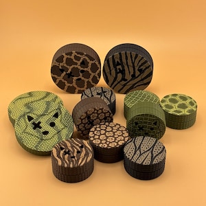 May include: A collection of circular, multi-layered herb grinders featuring various animal-inspired patterns including leopard, zebra, and snake skin prints. Several grinders display a stylized fox face cutout on the top surface.