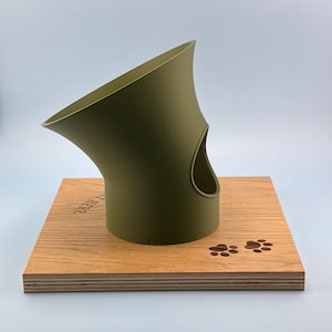 Pode incluir: Um objeto de plástico verde-oliva em forma de funil com uma abertura lateral, montado numa base quadrada de madeira. A base apresenta duas pegadas de pata incrustadas em madeira escura e o texto parcial "AT HERE" gravado na madeira clara.