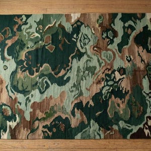 May include: Rectangular area rug with a camouflage pattern in shades of green, brown, and beige. The rug has a fringed edge and is placed on a wooden floor. The design features abstract shapes.