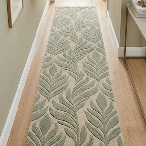 Puede incluir: Una alfombra larga y rectangular con un patrón de hojas repetido en tonos verdes y beige. La alfombra está colocada en un pasillo con suelo de madera y paredes claras. El diseño de hojas añade un toque natural al interior.