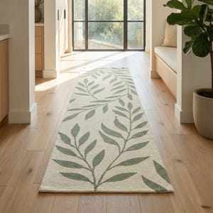 Puede incluir: Una alfombra larga y rectangular con un fondo color crema y un patrón repetido de hojas verde salvia estilizadas. La alfombra está colocada sobre un suelo de madera en un pasillo bien iluminado, con una gran ventana visible al fondo.