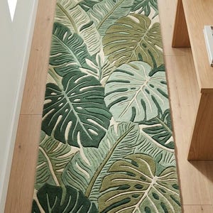 Puede incluir: Una alfombra larga y rectangular con un diseño de hojas tropicales en tonos verdes y crema. El patrón de la alfombra incluye grandes hojas de monstera y otro follaje tropical, creando un tema natural y botánico.