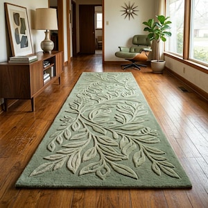 May include: A long, rectangular, sage green rug with a raised, botanical design. The rug features a central pattern of stylized leaves and branches in a lighter shade of green, creating a textured, dimensional effect. The rug is placed on a hardwood floor.