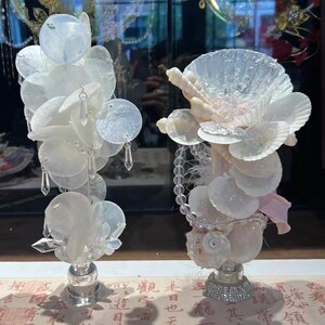 Könnte beinhalten: Zwei dekorative Muschelbildhauerarbeiten. Eine zeigt eine Ansammlung weißer, schillernder, kreisförmiger Muscheln mit hängenden Kristallakzenten. Die andere hat eine große, fächerförmige Muschel und kleinere Muscheln, mit einer Perlenkette und einer Strassbasis.