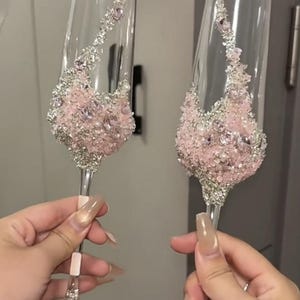 May include: Two clear champagne flutes with intricate crystal and pink bead embellishments. The stemware features a cascade of pink and silver crystals, creating a glamorous and elegant design. Perfect for celebrations and special occasions.