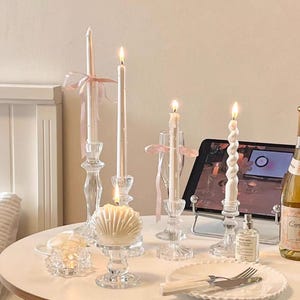 Könnte beinhalten: Eine Sammlung brennender Kerzen in Glashaltern und eine muschelförmige Kerze auf einem weißen Tisch. Die Kerzen haben verschiedene Formen und Größen, einige mit rosa Bändern. Eine Weinflasche und ein Tablet sind ebenfalls vorhanden.
