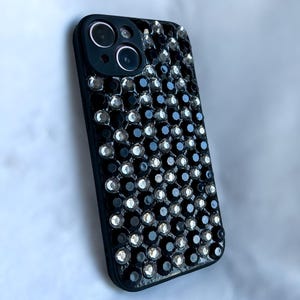 Peut inclure: Coque de smartphone noire ornée de petits strass noirs et argentés scintillants. L'étui comporte une ouverture pour l'appareil photo et est conçu pour un smartphone. Les strass sont disposés de manière dense, créant un effet chatoyant.