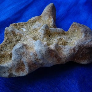 Pode incluir: Uma rocha de formato único e cor clara, com uma superfície áspera e texturizada. A rocha apresenta uma mistura de tons bege, castanho e cinza, com uma pequena protuberância pontiaguda. Repousa sobre um tecido azul vibrante, criando um contraste marcante.