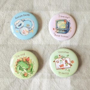 Könnte beinhalten: Vier runde Ansteckbuttons mit einzigartigen Designs. Ein Button ist hellblau mit dem Text "Autism Swag" und Kopfhörern. Ein weiterer ist rosa mit einem Retro-Computer und dem Text "I love my Puter friends". Ein dritter ist grün mit einem Dinosaurier und dem Text "I'm old". Der letzte ist grün mit einer Zeichnung und dem Text "Continue Drawing".