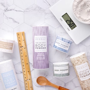 Puede incluir: Una vista cenital de varios productos de polvos corporales, incluyendo un tubo alto de lavanda y varios recipientes peque&ntilde;os. Tambi&eacute;n hay una regla de madera, una cuchara de madera y una balanza digital con un cuenco de polvo. Los productos est&aacute;n etiquetados con la marca y el aroma.