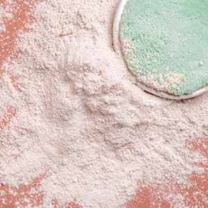 Puede incluir: Primer plano de un polvo beige claro esparcido sobre una superficie color melocot&oacute;n. Una almohadilla redonda, verde y esponjosa con un borde plateado es parcialmente visible, descansando sobre el polvo. La imagen tiene una apariencia suave y texturizada.
