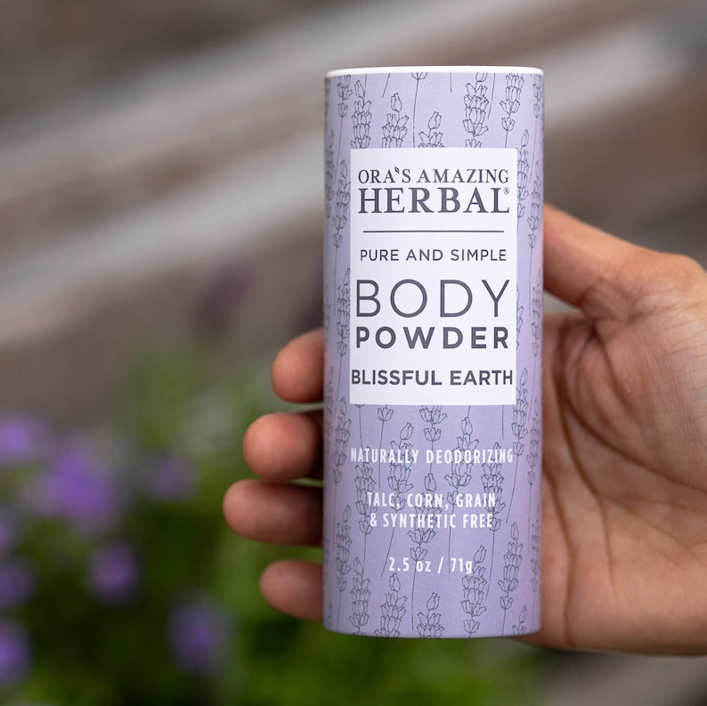 Puede incluir: Un envase cil&iacute;ndrico de polvos corporales con una etiqueta lavanda y blanca. La etiqueta dice "ORA'S AMAZING HERBAL BODY POWDER BLISSFUL EARTH". Contiene 71g de polvo.