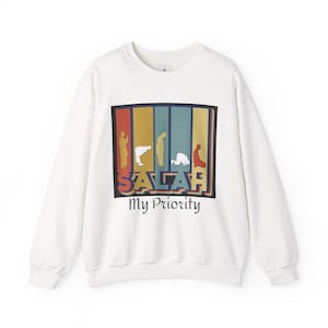 May include: White sweatshirt with a colorful graphic. The graphic features silhouettes of people in prayer, with the word "SALAH" and the phrase "My Priority" in a retro font. The design is set against a backdrop of vertical stripes in various colors.