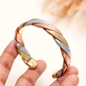 May include: A cuff bracelet featuring a triple-strand twisted design. The bracelet is constructed from three distinct metal bands in polished copper, brass, and silver tones, woven together to create a continuous, rounded, and open-ended circular form.