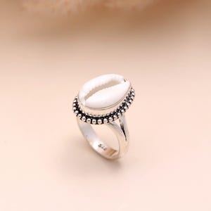 May include: A silver ring featuring a white seashell centerpiece. The seashell is oval-shaped and set within a beaded border. The ring band is a simple, polished silver. The ring is a piece of jewelry.