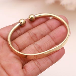 Op de afbeelding: Een goudkleurige manchetarmband met een afgerond, open ontwerp. De armband heeft twee kleine, bolvormige accenten aan de open uiteinden. Het gladde, gepolijste metaal reflecteert licht, waardoor een heldere, glanzende uitstraling ontstaat. Deze armband is een sieraad.