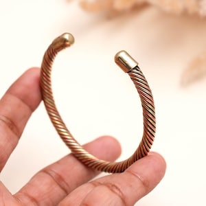May include: A cuff bracelet featuring a twisted design made of alternating strands of copper, brass, and silver-toned metal. The bracelet has a circular opening and is finished with smooth, rounded metal caps at both ends of the twisted band.