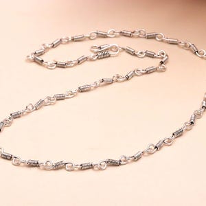 Puede incluir: Un collar plateado con un diseño único. El collar presenta una serie de pequeños segmentos de alambre enrollado conectados por eslabones ovalados. Un cierre es visible en el centro. La joya se muestra sobre un fondo beige claro.