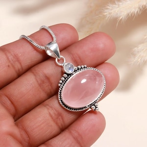 Puede incluir: Un collar con colgante de plata con una gran gema ovalada de cuarzo rosa. El colgante tiene una piedra lunar más pequeña, redonda e iridiscente y un borde decorativo. La cadena es de plata delicada y texturizada.
