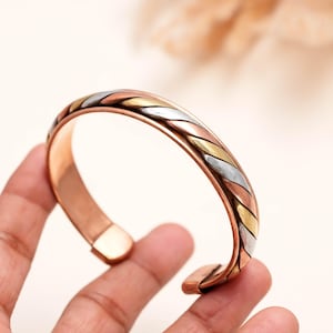 May include: A cuff bracelet featuring a tri-color design with twisted strands of copper, brass, and silver-toned metal. The open-ended band has a smooth, polished finish on the interior and a braided, multi-metal texture on the exterior surface.