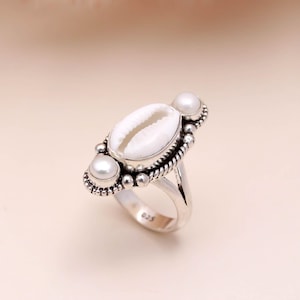 May include: A silver ring featuring a white cowrie shell center stone, flanked by two white pearl accents. The ring has a detailed, textured setting with a rope-like border and small beaded accents. The ring is on a light background.