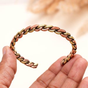May include: A cuff bracelet featuring a braided design made of alternating copper and brass metal links. The bracelet has an open-ended circular shape with a textured, metallic finish that displays the distinct reddish-brown and golden hues.