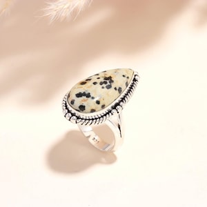May include: A silver ring featuring a large, teardrop-shaped stone with a cream-colored base and black spots. The stone is set in a decorative, rope-like bezel. The ring band is silver and stamped with "925".