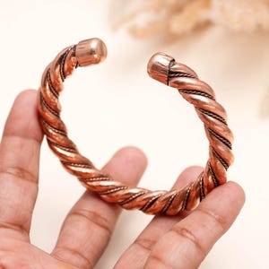 Op de afbeelding: Een koperen armband met een gedraaid touwontwerp en een gepolijste afwerking. De armband heeft een opening in het midden met afgeronde, gladde uiteinden. Het metalen oppervlak vertoont over het hele sieraad een warme, roodbruine kleur.