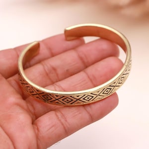 May include: A gold-colored cuff bracelet with a geometric pattern of small diamonds etched around the band. The open-ended bracelet is shown on a hand, showcasing its design and style. This accessory is suitable for everyday wear.