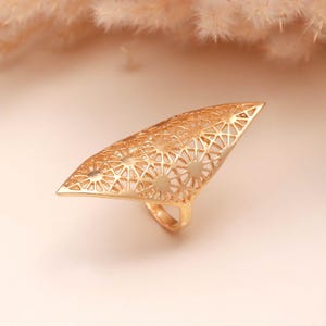 May include: Gold-colored statement ring with an openwork design. The ring features a large, triangular top with a geometric pattern of circles and lines. The ring's band is a simple, smooth gold. The ring is a bold piece of jewelry.