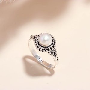 May include: A silver ring with a large, round, white pearl at its centre. The pearl is set in an ornate, detailed setting with a rope-like border and decorative features. The ring has a polished finish and a classic, elegant design.