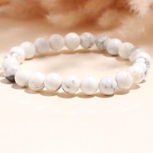 Puede incluir: Pulsera de cuentas de mármol blanco. La pulsera presenta cuentas redondas y pulidas con una mezcla de patrones blancos sólidos y grises veteados. Las cuentas están ensartadas para formar una forma circular, adecuada para usar en la muñeca. La pulsera se muestra sobre una superficie clara.