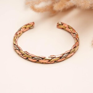 May include: A cuff bracelet featuring a braided design composed of three distinct metallic strands in copper, brass, and silver tones. The open-ended circular band displays a polished finish with interlocking segments that create a textured appearance.