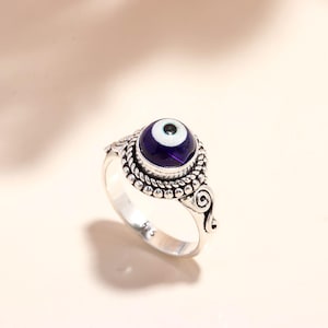 May include: A silver ring featuring a large, round, dark blue eye-shaped stone with a white iris and black pupil. The ring has an ornate, textured band with a beaded design and scrollwork details. The ring is a piece of jewelry.