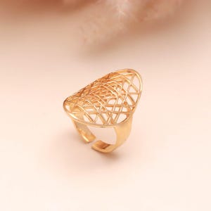 May include: A gold-colored ring with an open, geometric design. The ring features an elongated, oval-shaped top with a lattice pattern. The band is adjustable, with an open design at the bottom. The ring is set against a soft, neutral background.